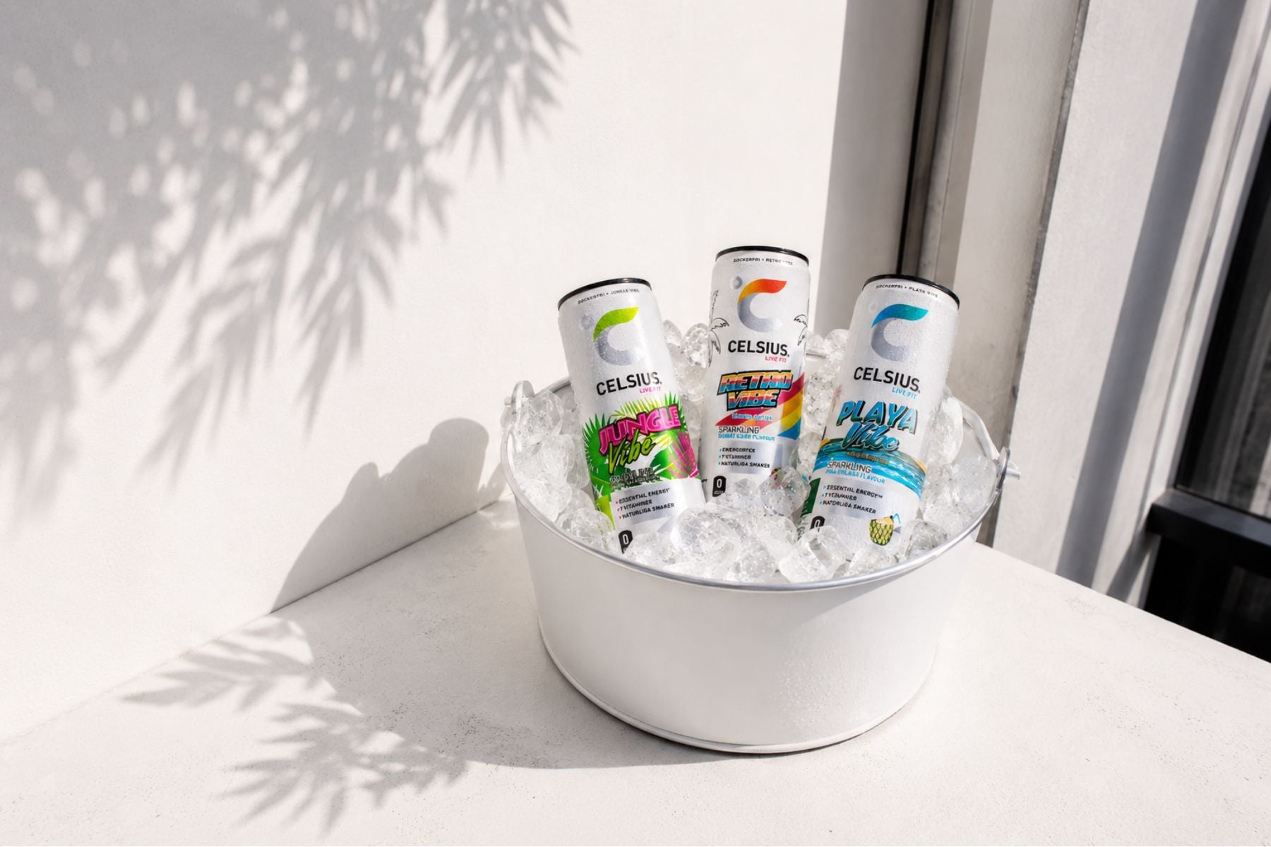 Three cans of Celsius energy drinks in a bowl on a light surface with a window in the background.