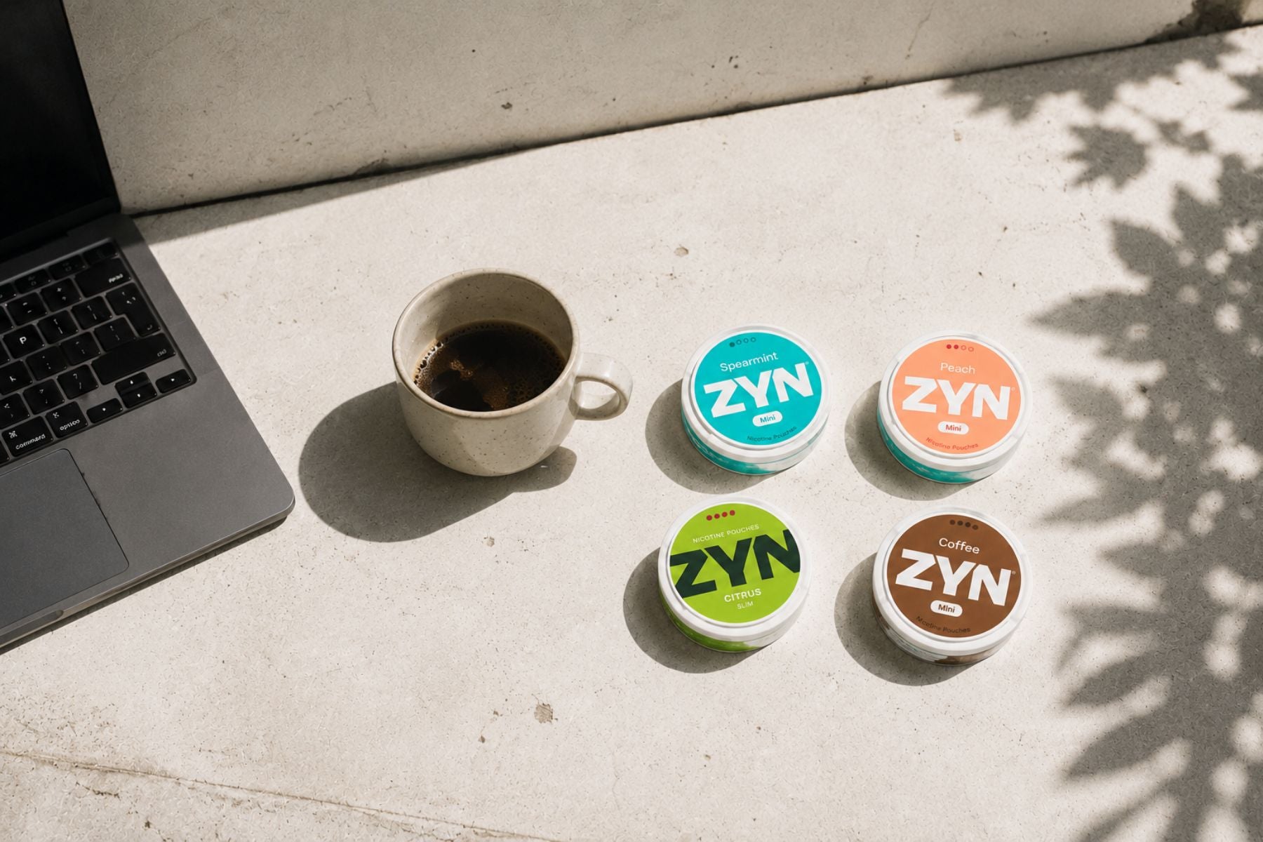 ZYN Nicotine Pouches on a concrete surface next to a cup of coffee and a laptop.