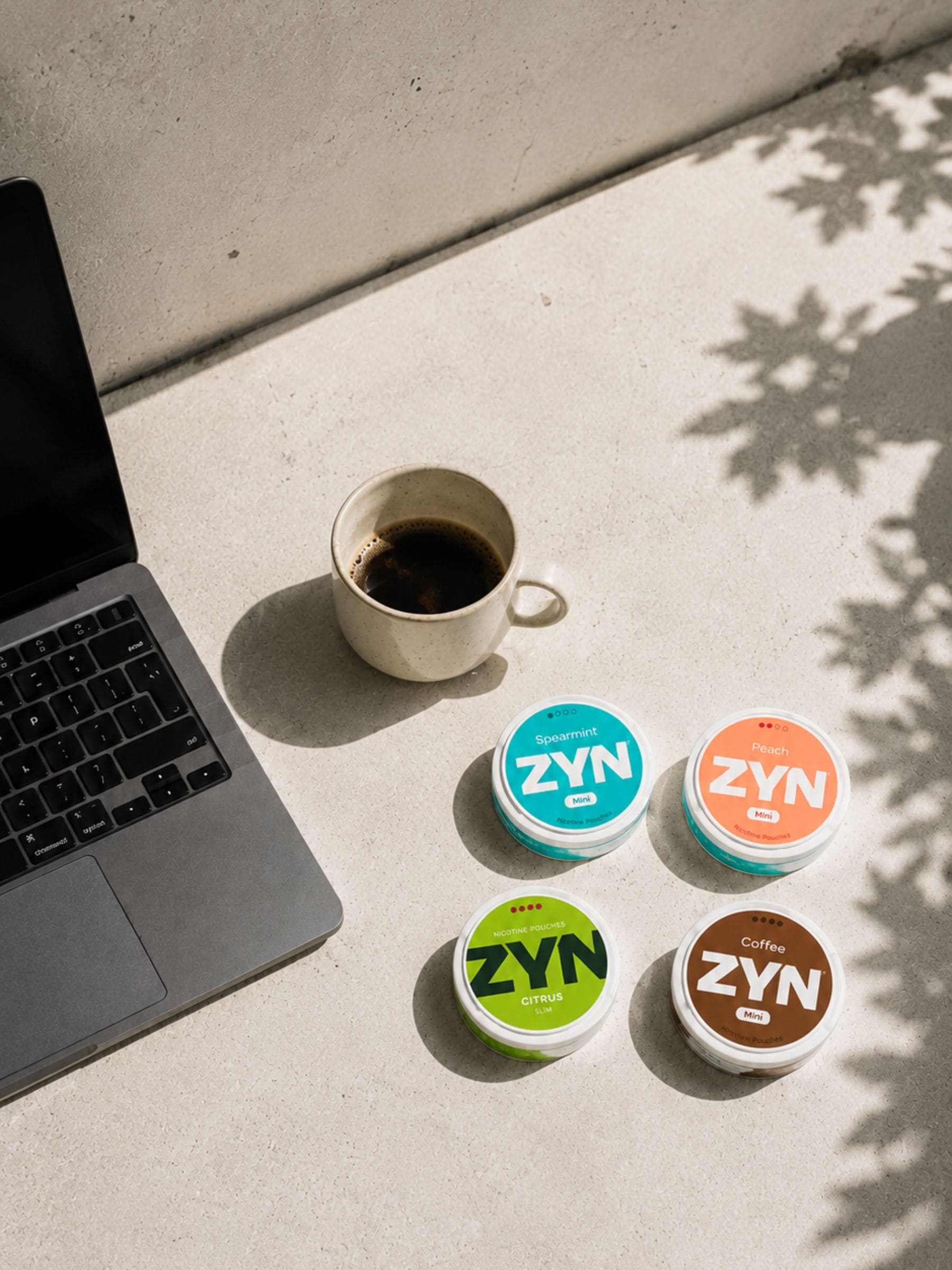 ZYN Nicotine Pouches on a concrete surface next to a cup of coffee and a laptop.